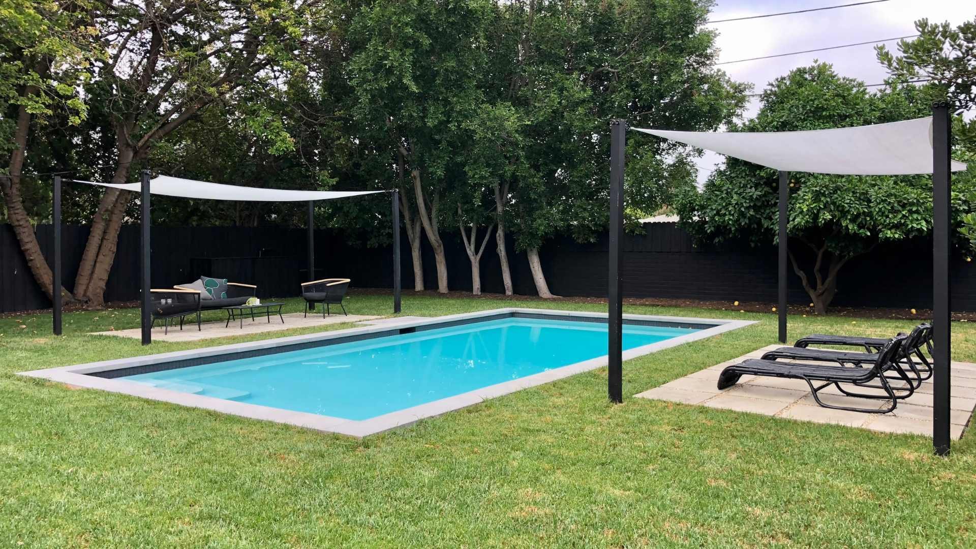 Rectangular pool with shade sails in a modern backyard, featuring a lush green lawn and black fence.