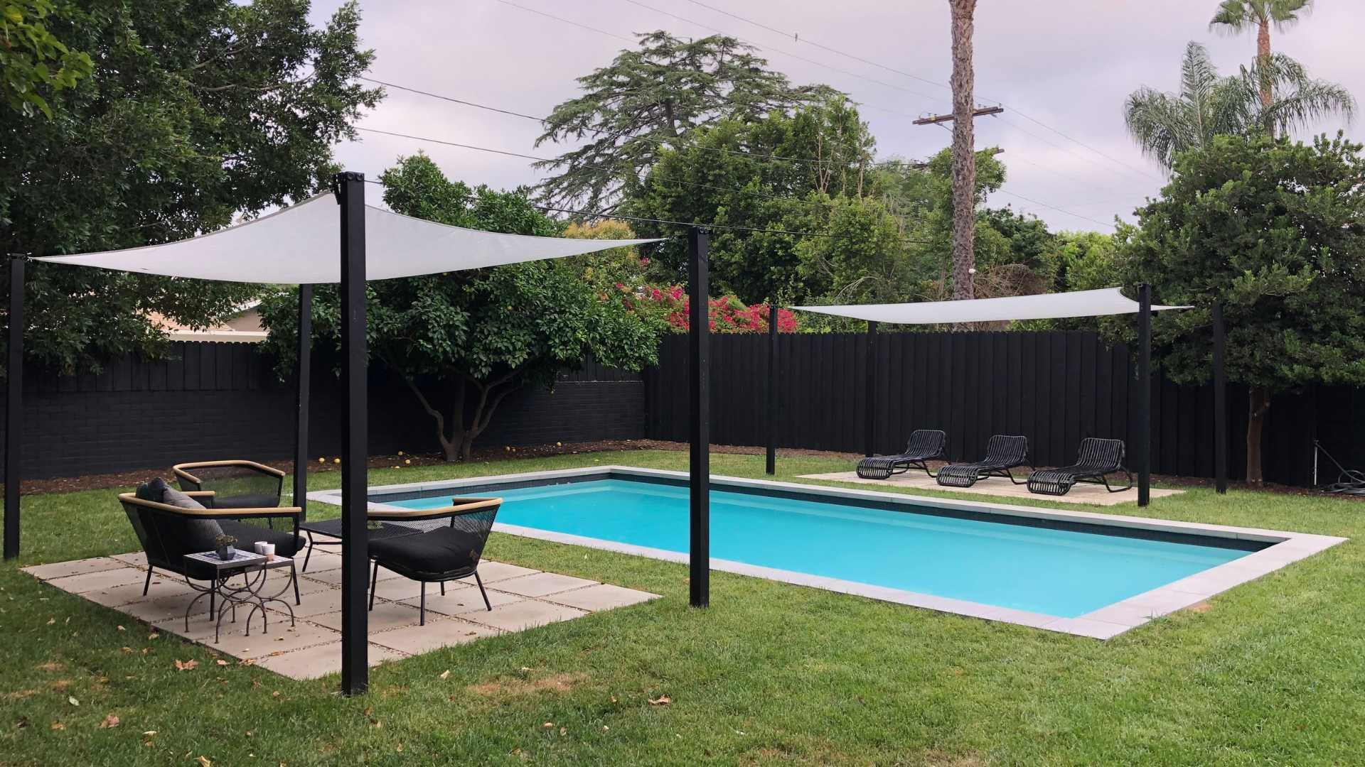 Wide shot of a rectangular pool with shade sails, set in a grassy backyard surrounded by mature trees and black fencing.