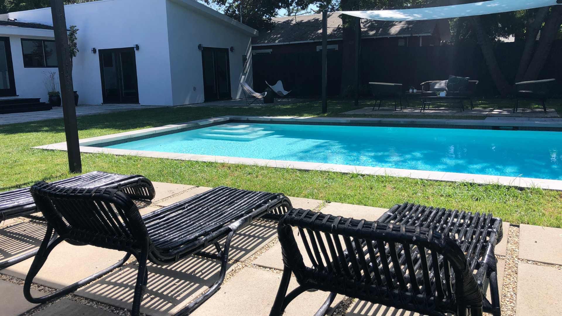 Side view of a rectangular pool with lounge chairs and shade sails, set in a grassy backyard with modern white buildings.