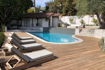 A curved pool with a wooden deck and lounge chairs next to a charming house with a red-tiled roof surrounded by trees and lush landscaping.