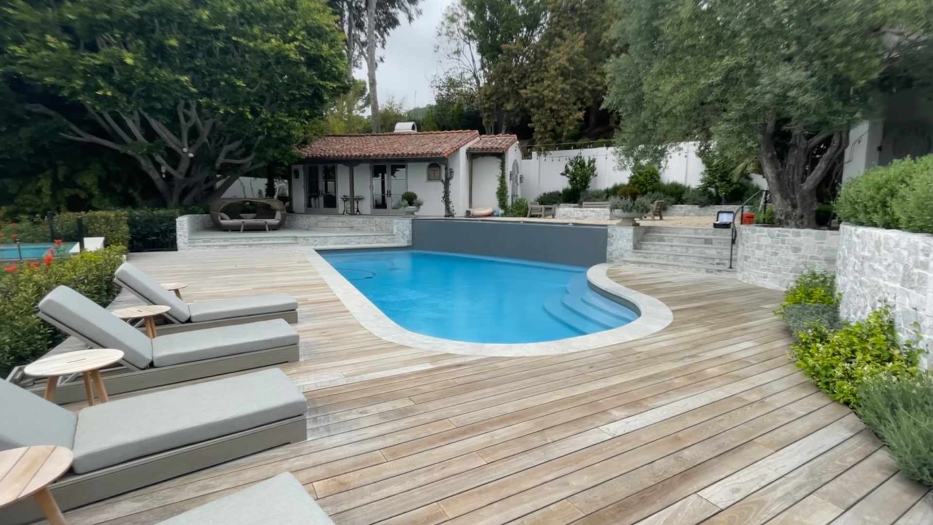 A curved pool with a wooden deck and lounge chairs next to a charming house with a red-tiled roof surrounded by trees and lush landscaping.