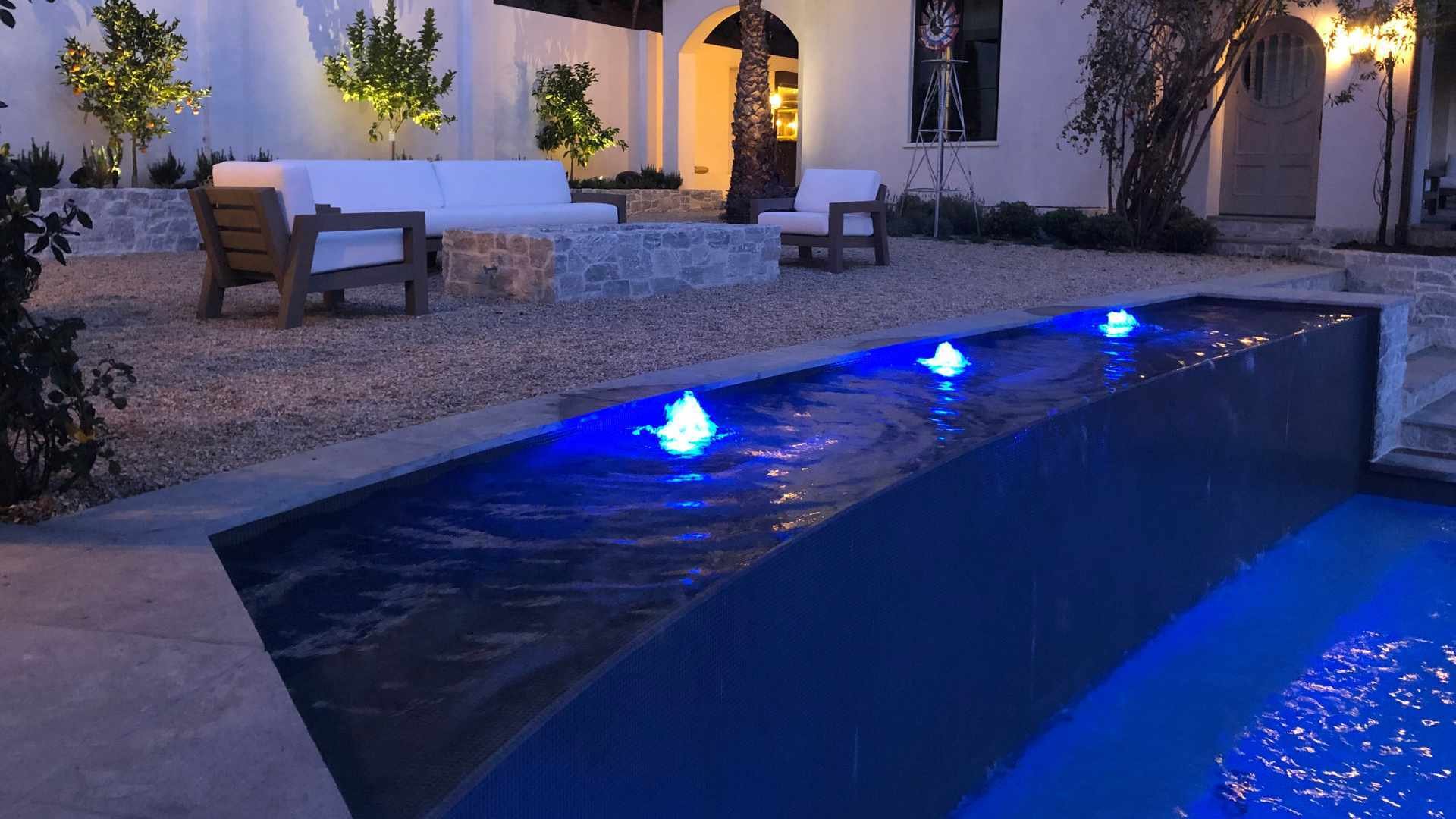 Illuminated pool at night with lights, next to a gravel seating area with string lights and palm trees.