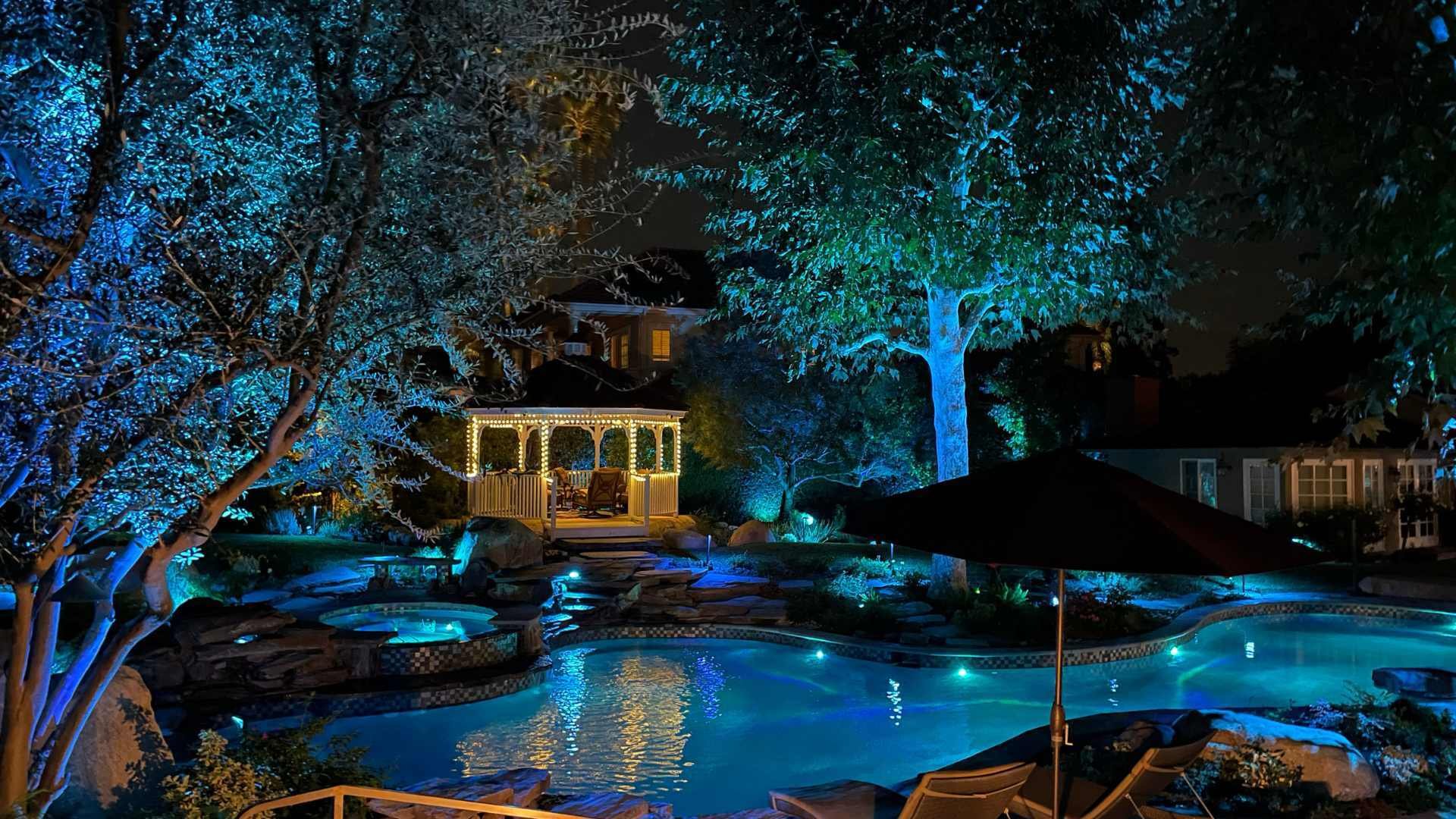 A nighttime view of a pool and spa area illuminated by landscape lighting and a gazebo in the background.