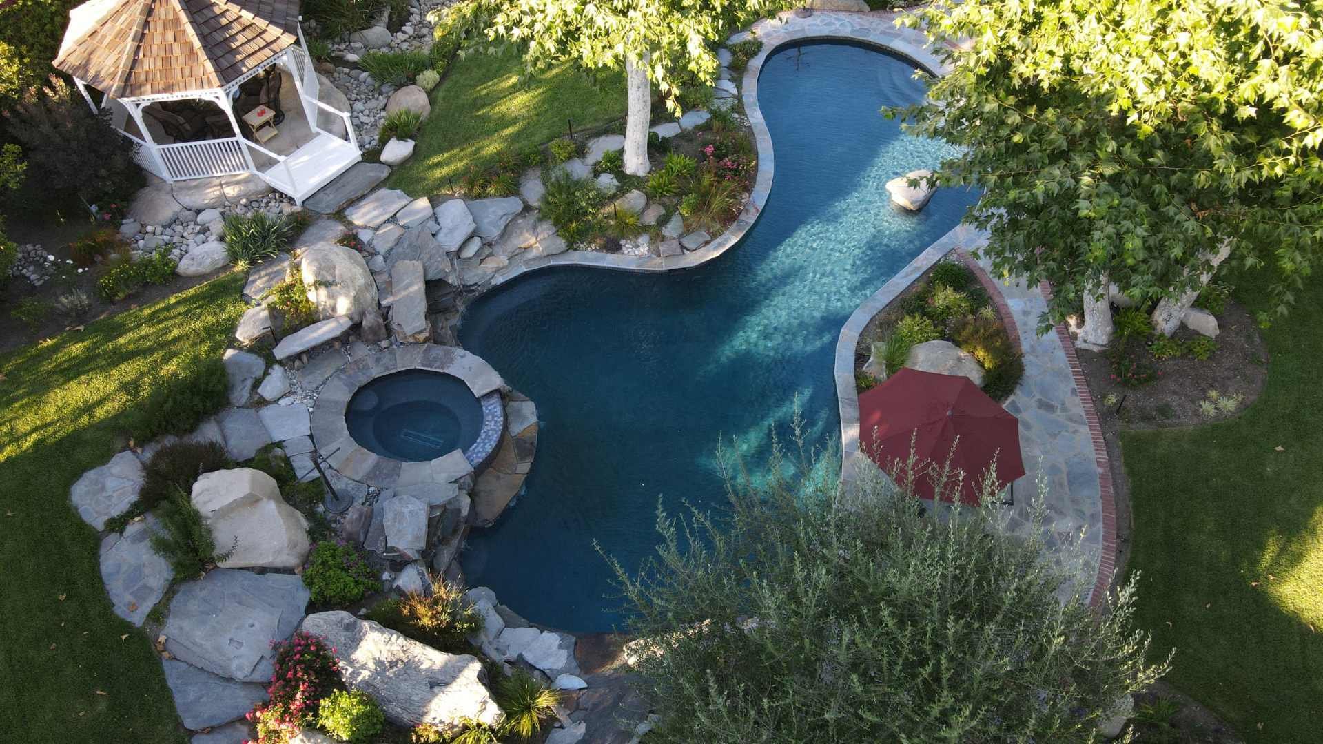 Aerial view of a uniquely shaped pool with a raised spa, natural stone landscaping, and a gazebo.