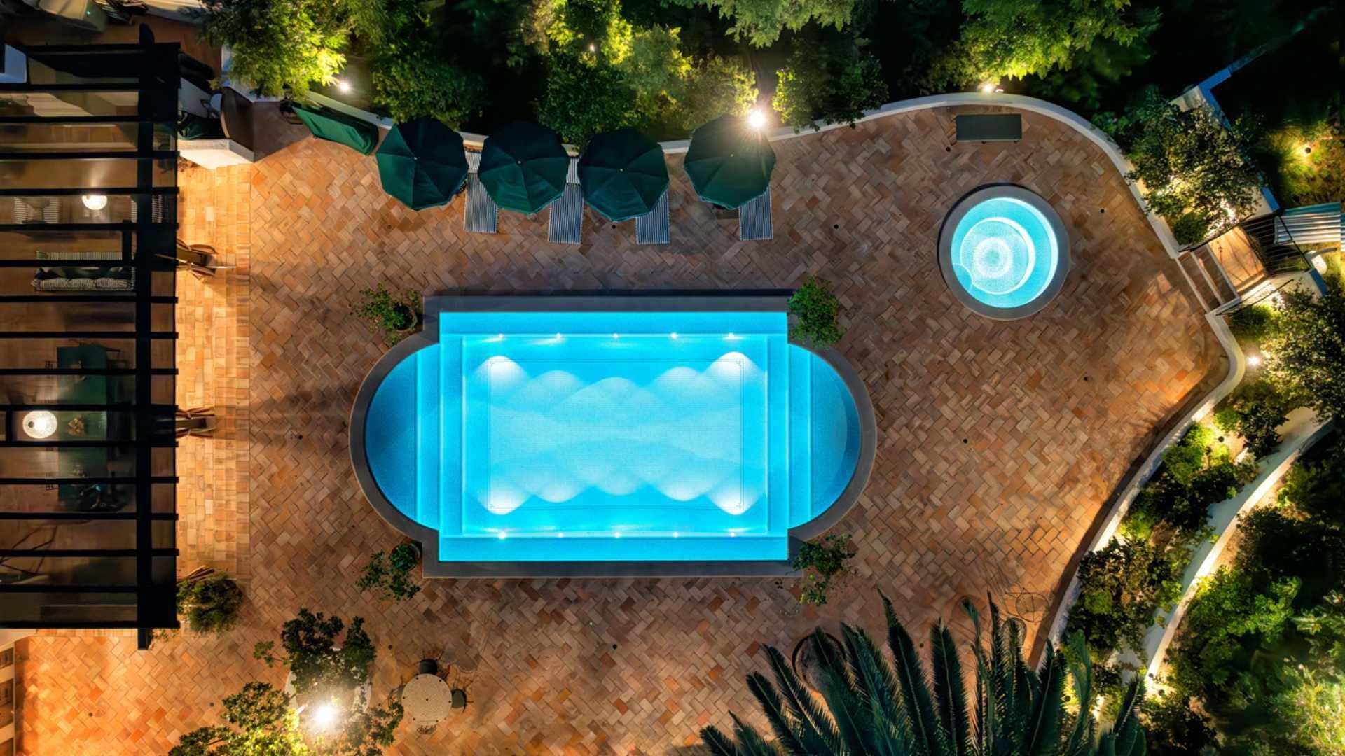 Arial view of a glowing, fully tiled pool at night, framed by terracotta tiles and potted plants, illuminated by LED lights.