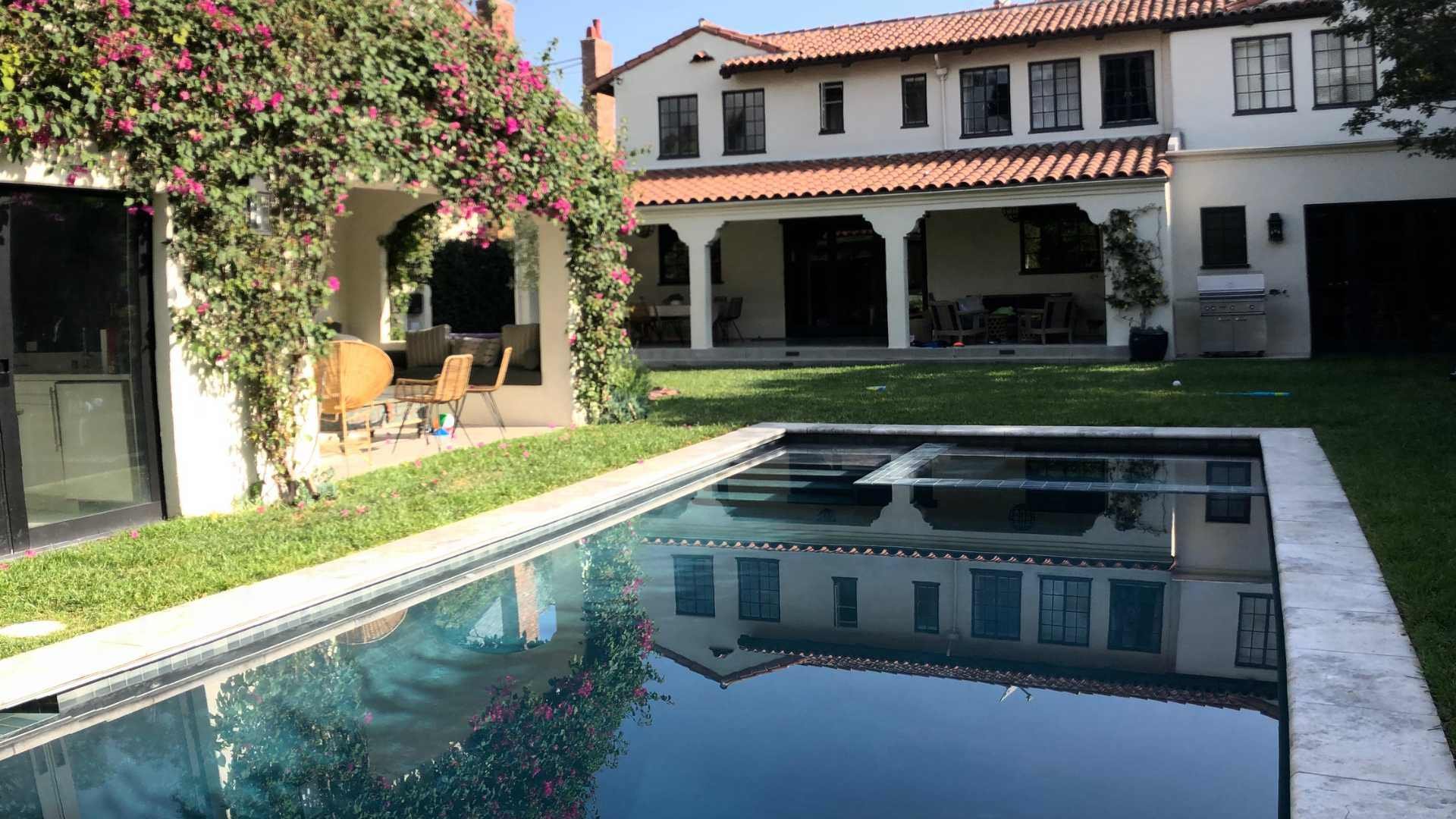 A tranquil pool in a Mediterranean-style backyard featuring a patio covered with blooming bougainvillea and a view of the house in the background.