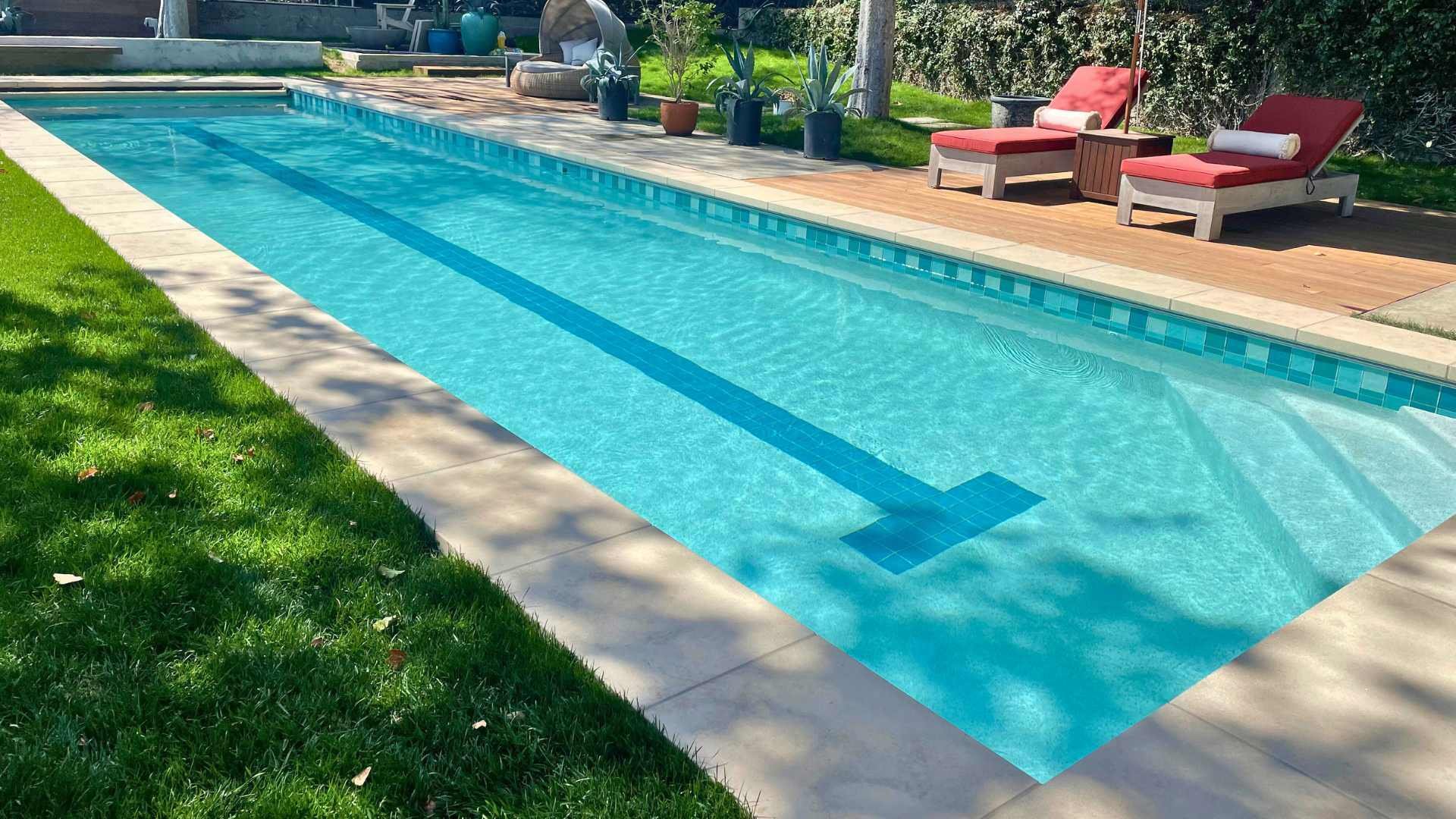 A rectangular pool with blue tile featuring a central lane marker, bordered by a green lawn, wooden deck, and shaded by trees.