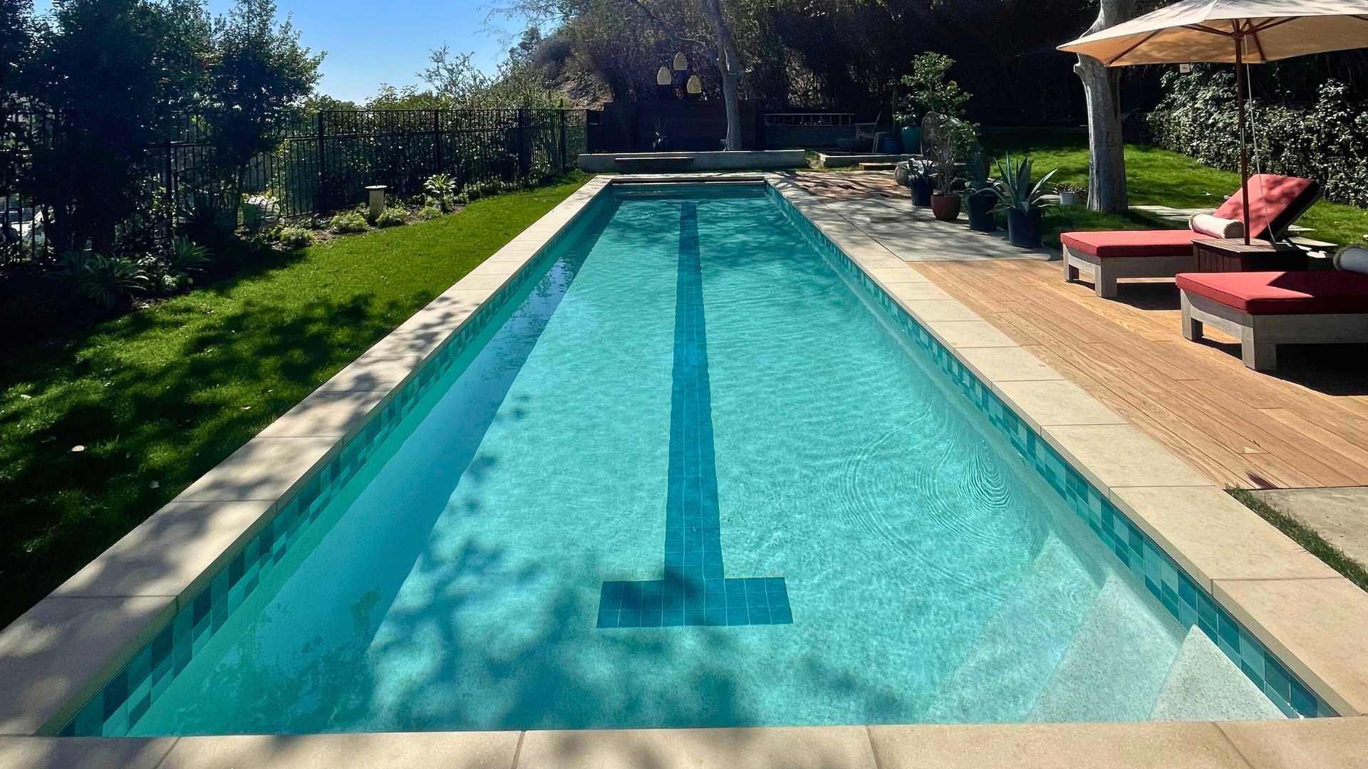 Long, narrow lap pool with a central lane marker, bordered by trees and lounge chairs, under a bright, sunny sky.