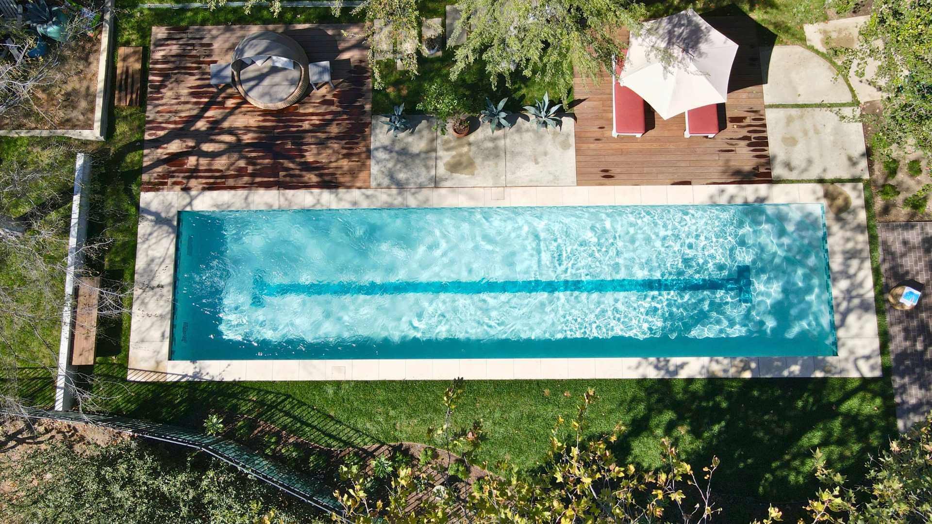 A lap pool with a central lane marker, surrounded by a wooden deck, lounge chairs, and lush greenery, as seen from above.