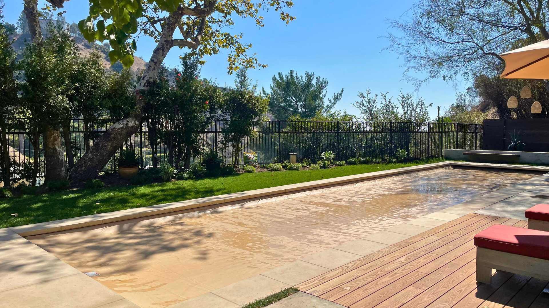Closed electrical cover on top of a rectangular lap pool surrounded by trees, a grassy lawn, and lounge chairs on a sunny day.