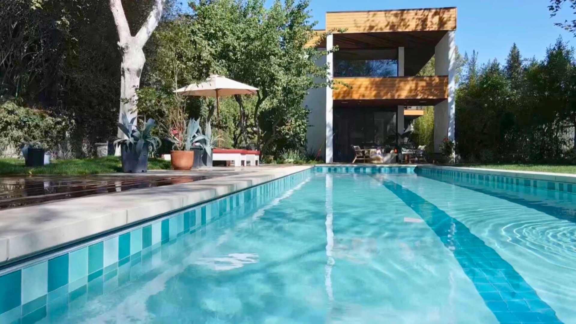A modern pool with a central lane marker surrounded by a wooden deck, lush greenery, and a contemporary house in the background.