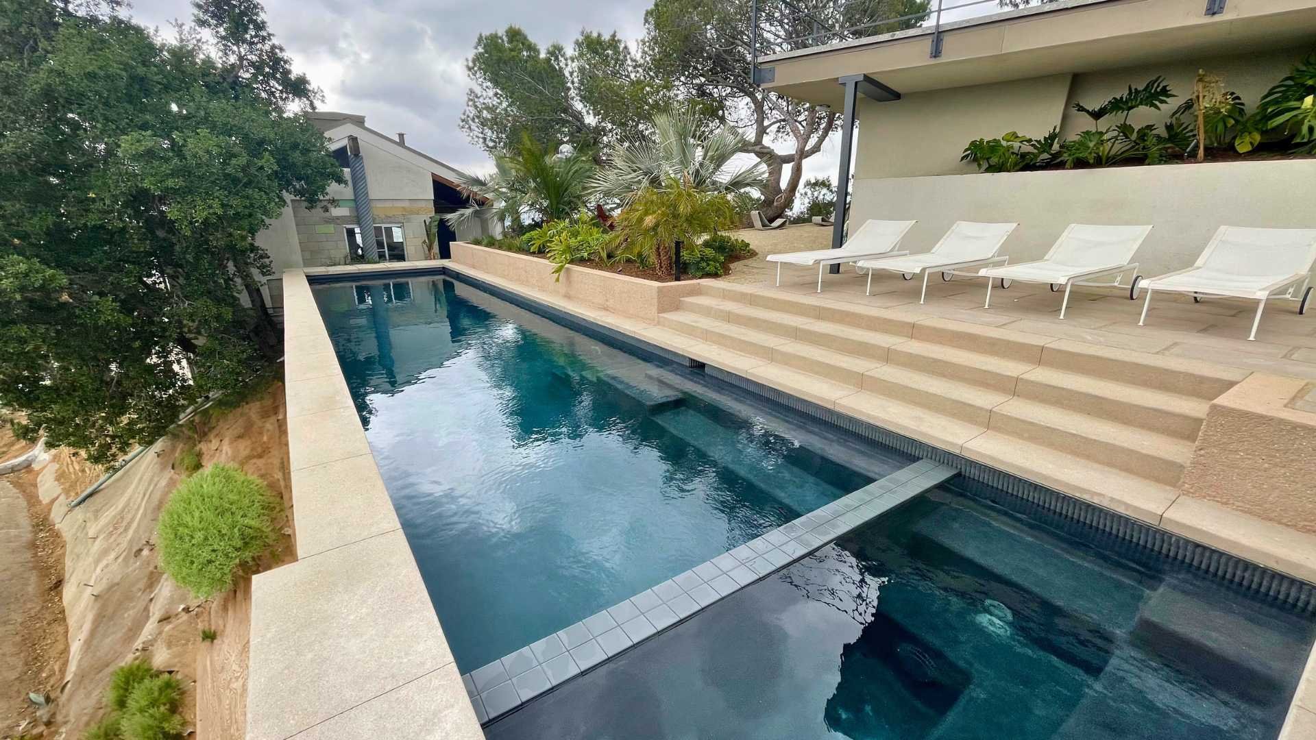 Sleek lap pool with steps leading to a deck, bordered by lounge chairs on a cloudy day.