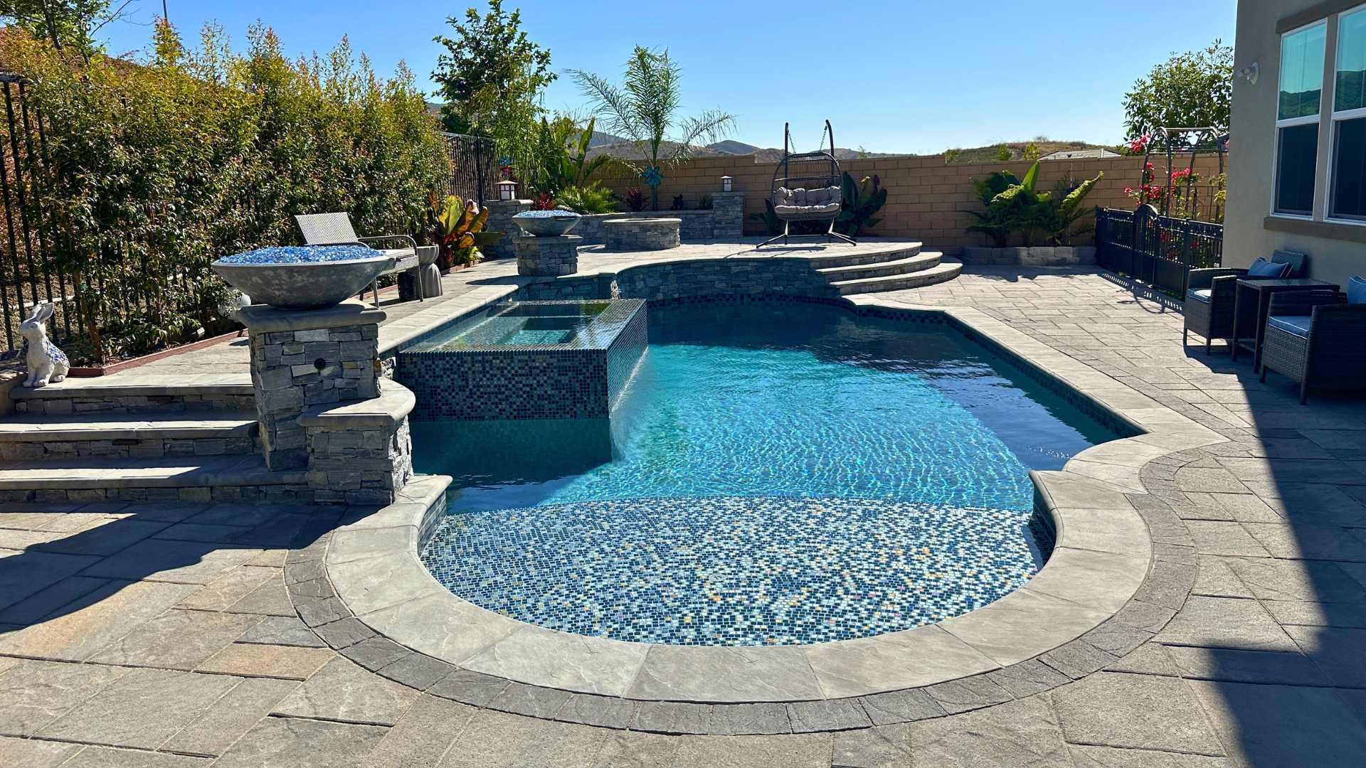 A luxurious backyard pool with a built-in hot tub, stone steps, and a modern patio area surrounded by greenery and outdoor seating