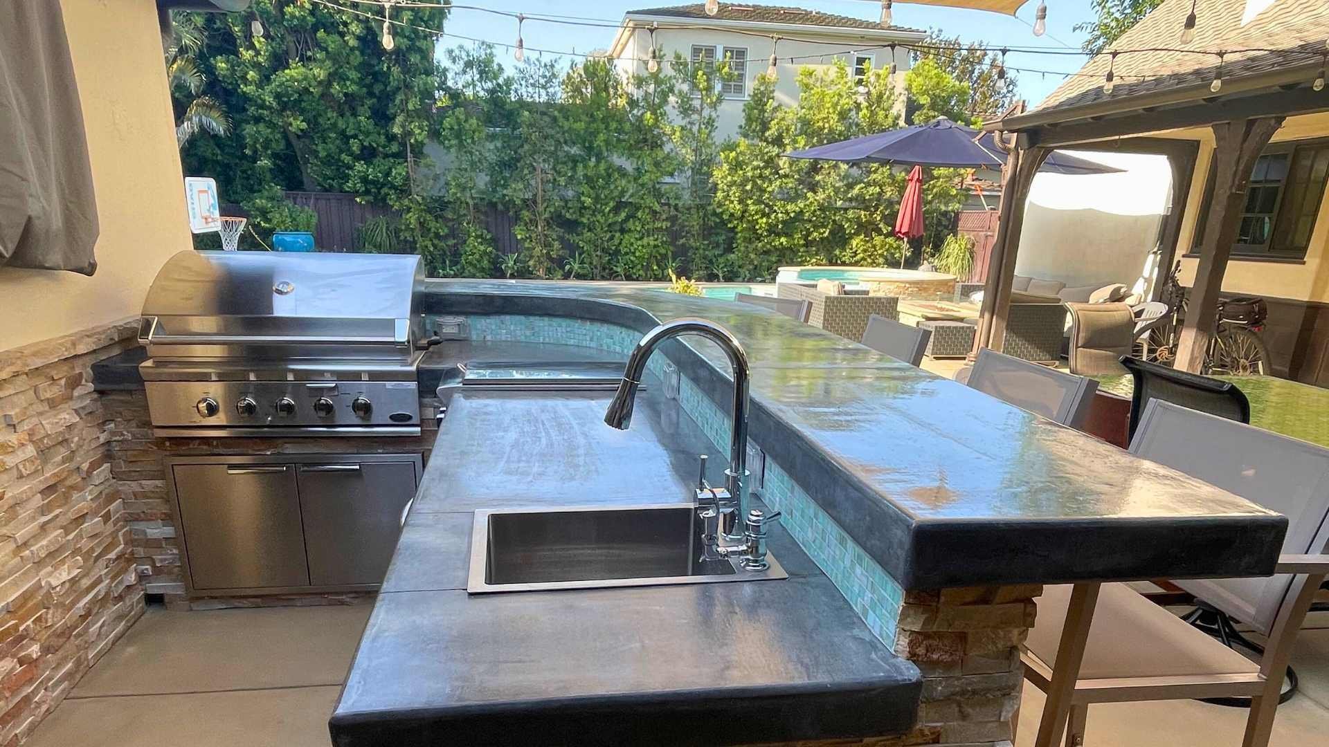 Outdoor kitchen with a built-in grill, sink, and stone counters, located near a pool in a modern backyard.
