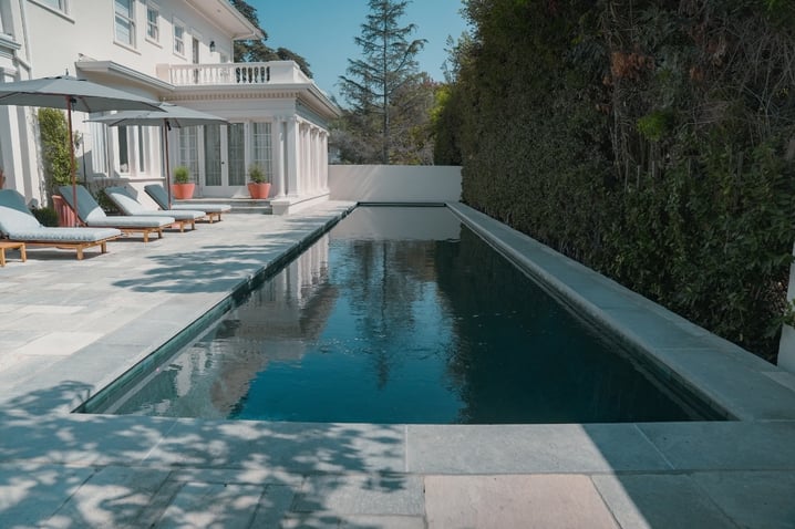 A long, narrow, dark color pool beside a white house with lounge chairs under umbrellas, bordered by tall hedges.