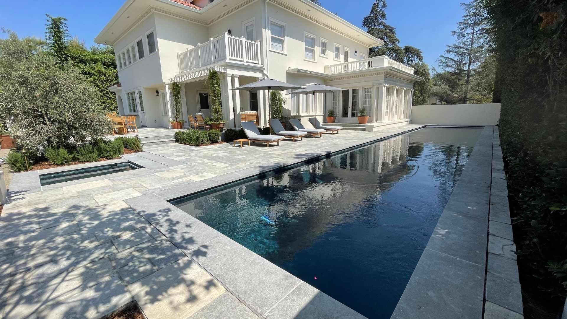 A long, narrow pool next to a white house, with lounge chairs and umbrellas on the stone patio, bordered by greenery.