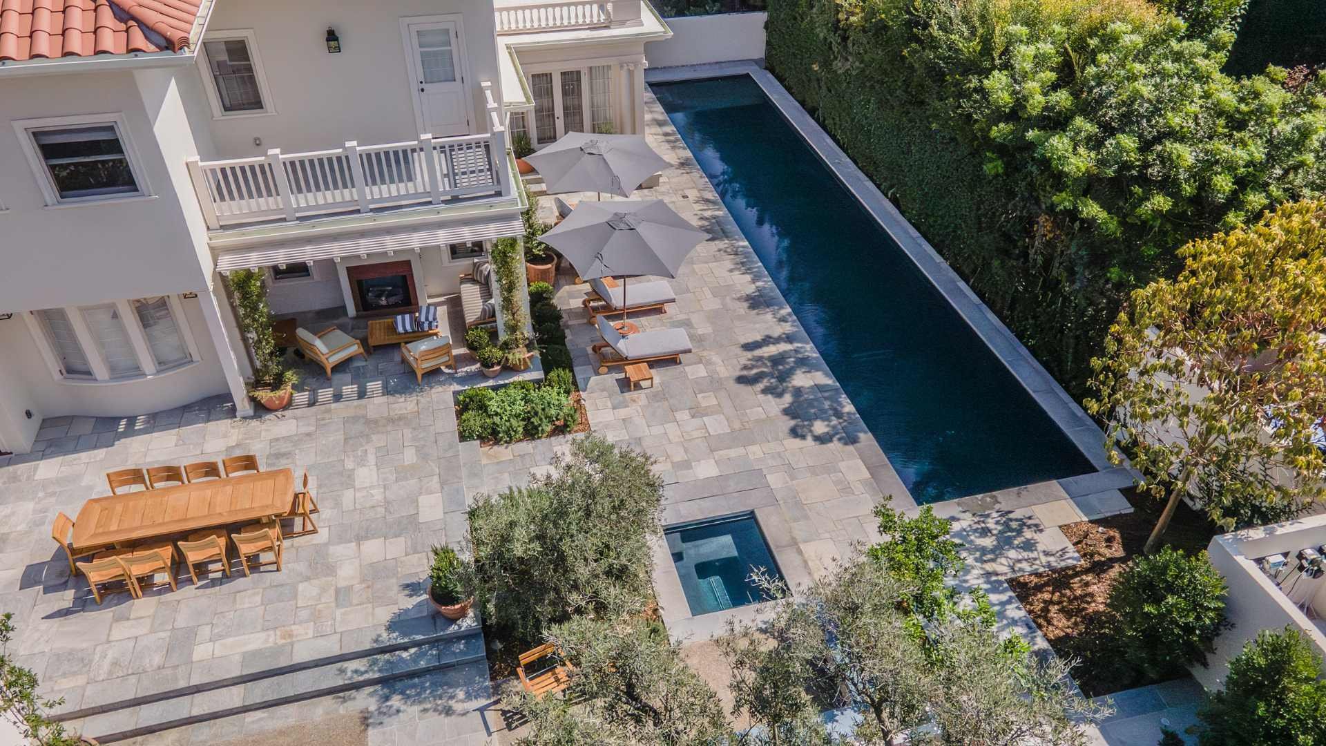 Aerial view of a spacious patio featuring a long, rectangular pool lined with lounge chairs and umbrellas, a dining table, and a hot tub.