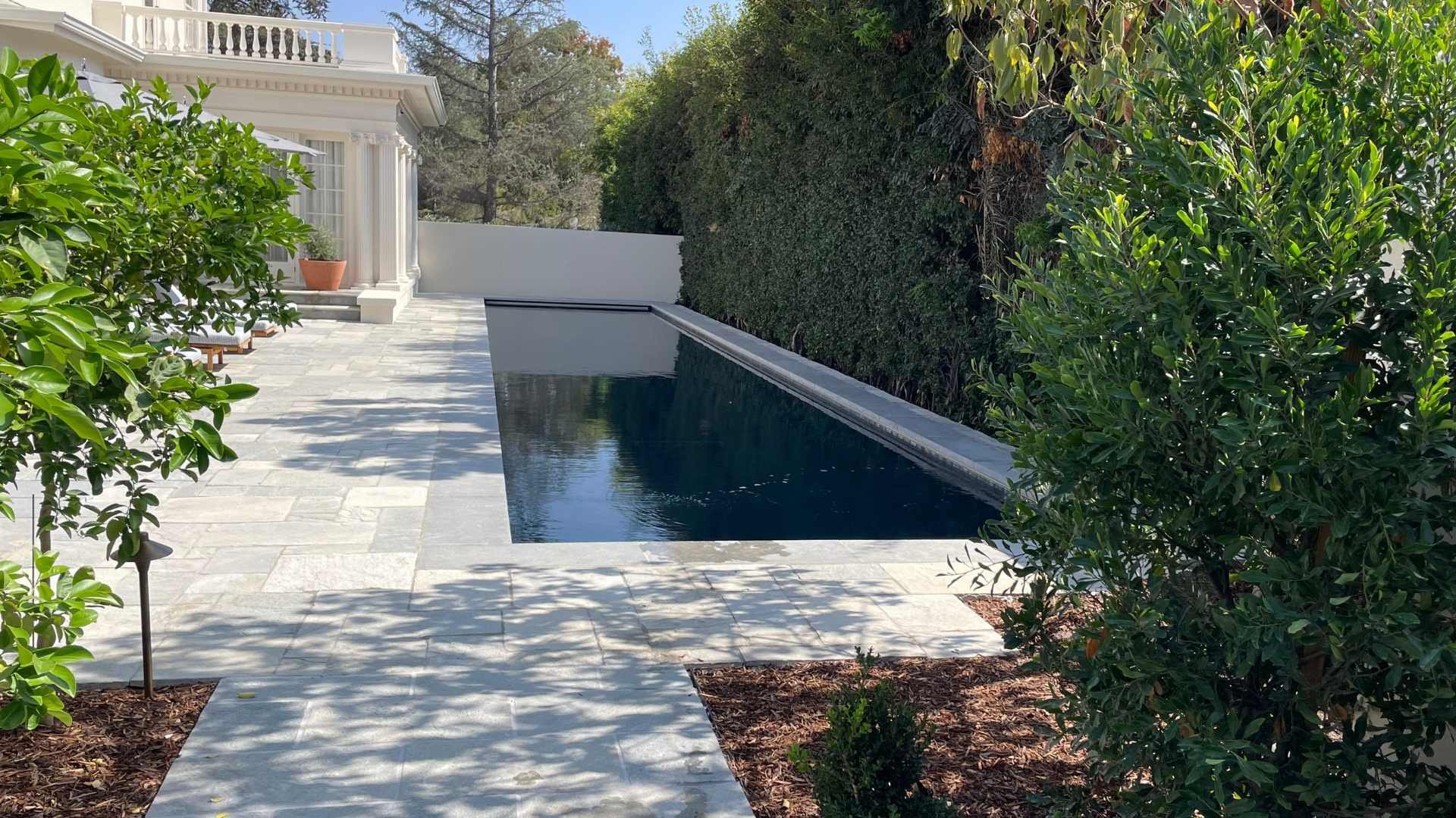 A long, narrow, dark color pool beside a white house with lounge chairs under umbrellas, bordered by tall hedges.