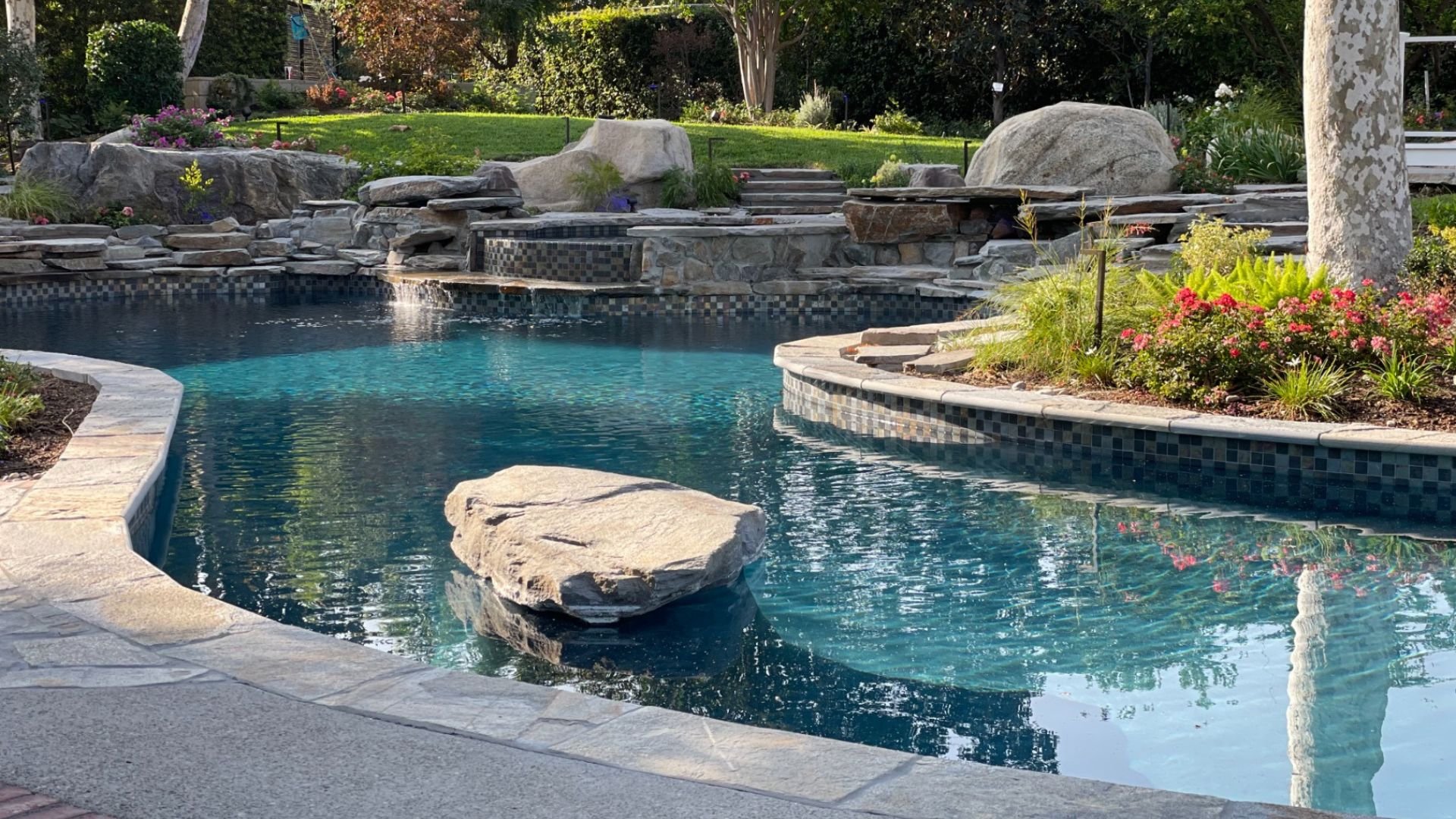 Lagoon-style pool & spa with natural rock waterfalls, radiant Lightstream glass tiles, and stunning blue ocean Primera stone finish.