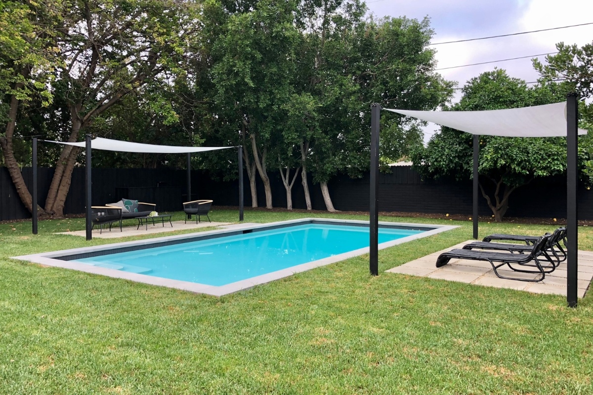 Rectangular pool with shade sails in a modern backyard, featuring a lush green lawn and black fence.