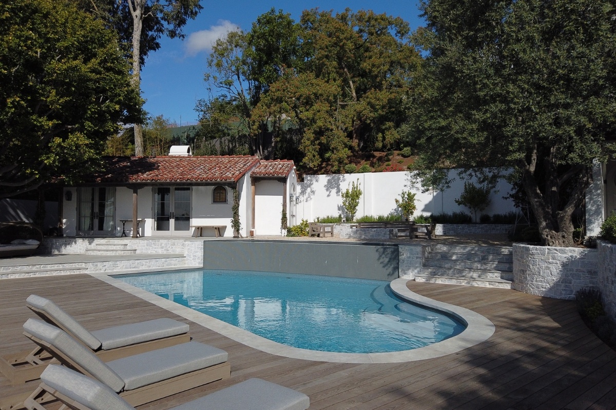 A curved swimming pool surrounded by a wooden deck with lounge chairs, set against a backdrop of a white house with red-tiled roofs and lush greenery.