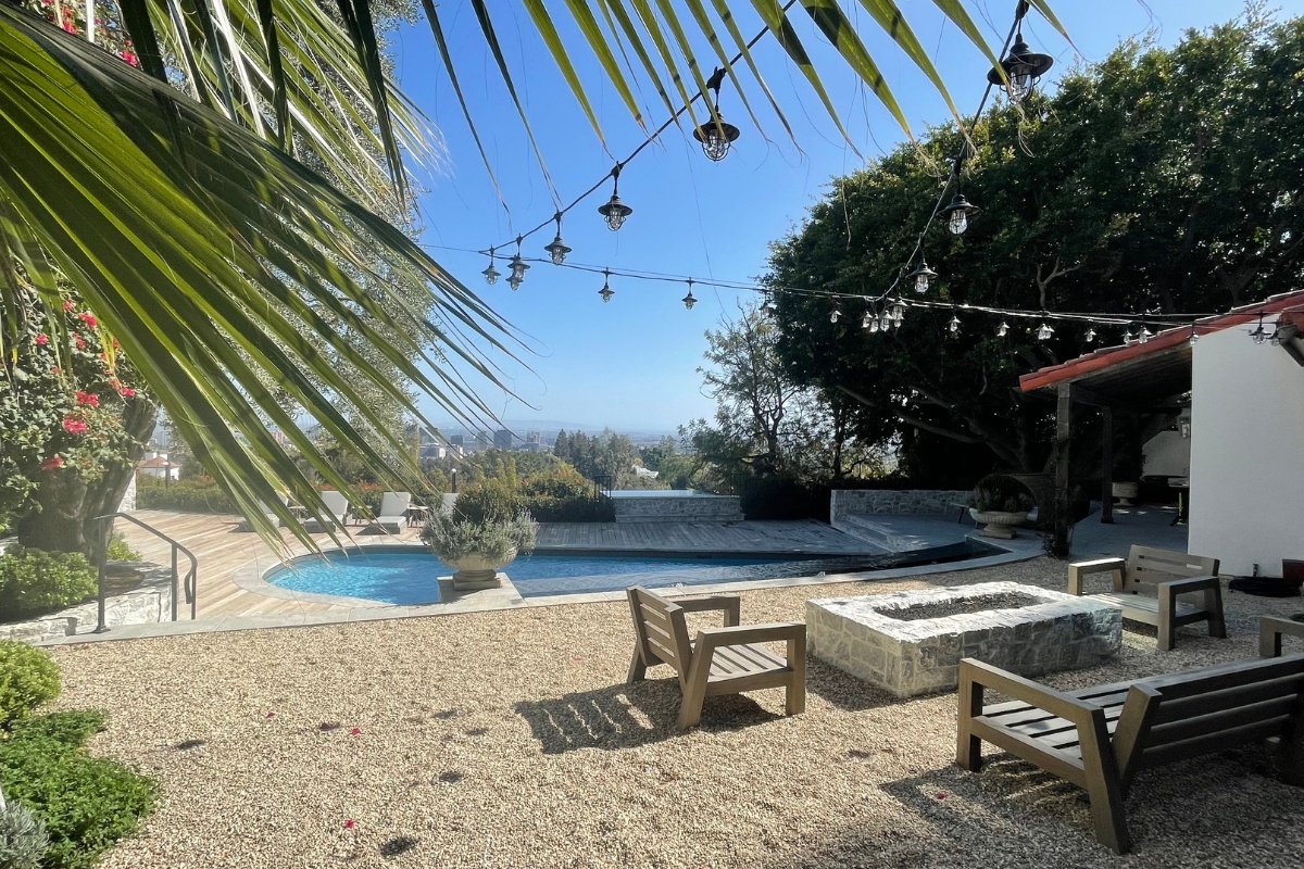 Gravel patio with string lights, wooden benches around a stone fire pit, and a view of a pool and wood deck