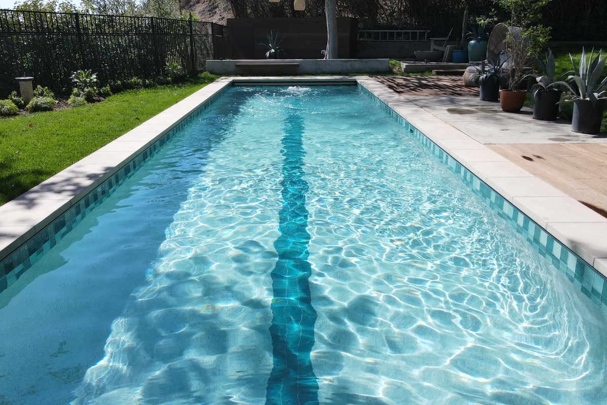 Close-up view of a lap pool with a lane marker, bordered by a wooden deck, plants, and a green lawn.