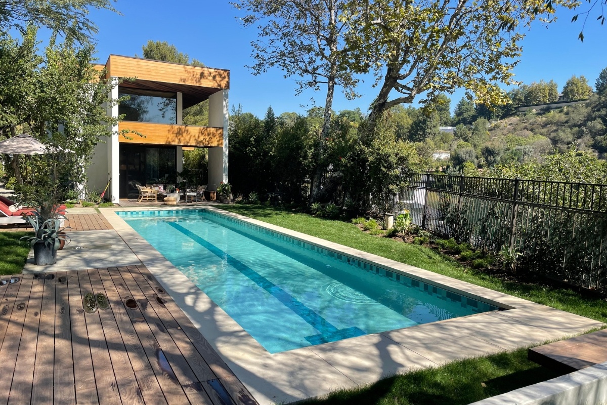 A modern pool with a central lane marker surrounded by a wooden deck, lush greenery, and a contemporary house in the background.