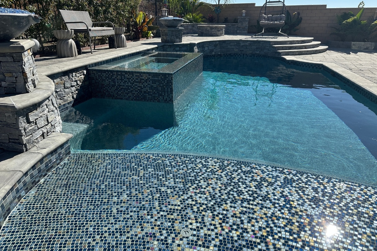 A close-up of a modern backyard pool featuring a mosaic tile design, integrated spa, and surrounding patio with elegant seating.