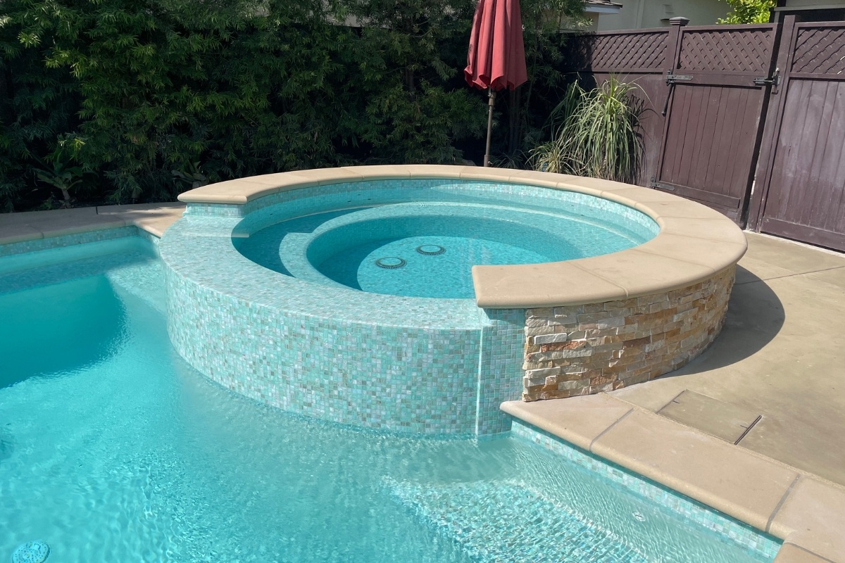 Close-up of a raised spa with light blue glass tile connected to a pool in a private backyard.