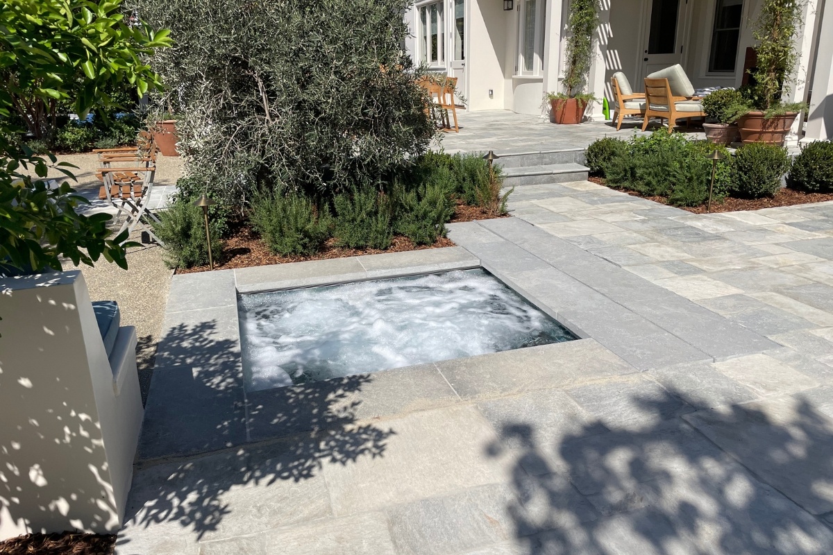 Sunken hot tub with bubbling water, surrounded by a stone patio and lush greenery, next to outdoor seating areas.