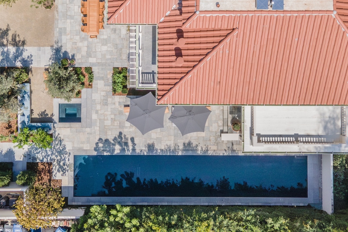 Aerial view of a long, rectangular pool and patio next to a red-roofed house with outdoor seating areas and landscaping.